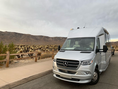 An Airstream rv rental parked roadside near a Cholla Cactus Garden with desert mountains under a cloudy sky.