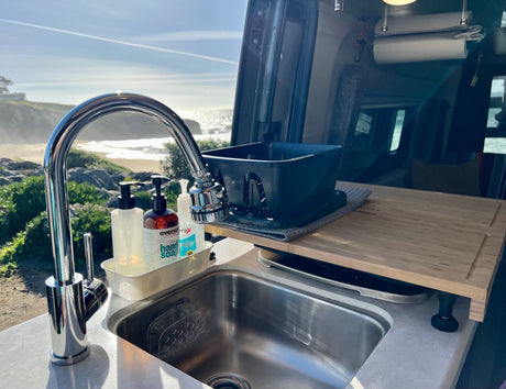 RV rental's clean kitchen sink area with soaps & dish rack, offering a stunning beach and ocean view from inside.