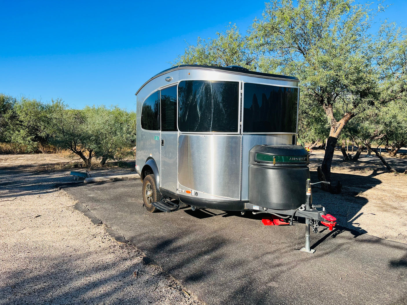 Silver Airstream Basecamp travel trailer parked on an asphalt pad, ready for your RV rental adventure among the trees.