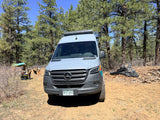 Outdoorsy rv rental Mercedes Sprinter camper van parked on a dirt road in a sunny pine forest with camping gear.