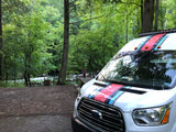 White Ford Transit rv rental, styled with stripes, at a scenic riverside campsite surrounded by lush green trees.
