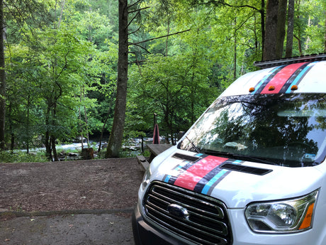 White Ford Transit rv rental, styled with stripes, at a scenic riverside campsite surrounded by lush green trees.