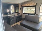 Modern **RV rental** living space: TV, stereo, Dometic AC/heater, and leatherette sofa overlooking a campsite.