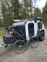 A compact, rugged rv rental camper trailer with multiple bikes is parked on gravel amidst tall pine trees.