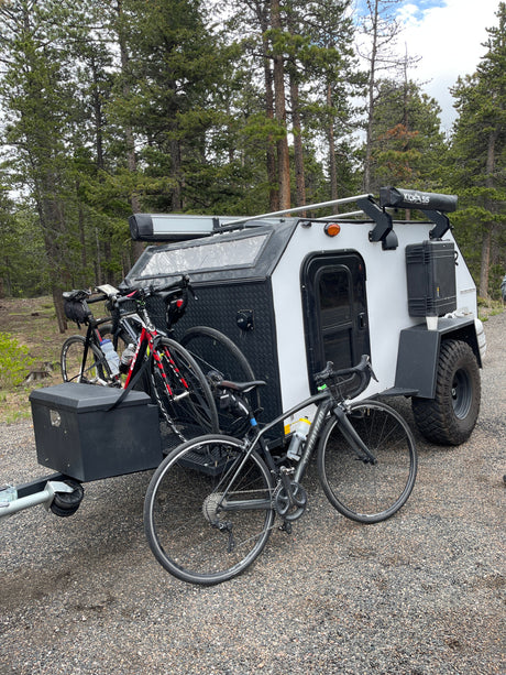 A compact, rugged rv rental camper trailer with multiple bikes is parked on gravel amidst tall pine trees.