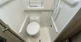 Compact wet bath in an RV rental, featuring a toilet, shower area with a drain, and an overhead storage shelf.