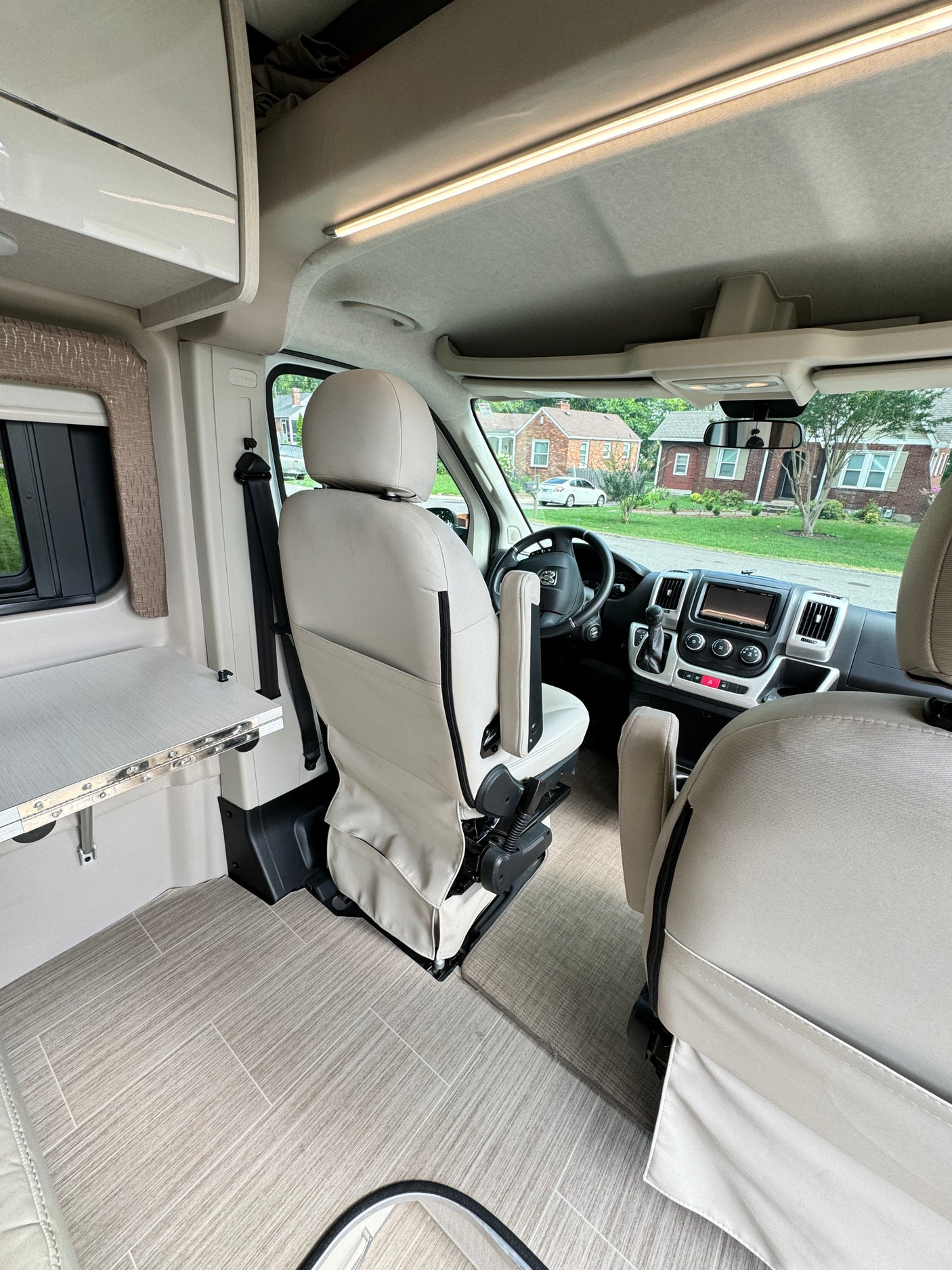 Bright RV rental interior view from rear: light swivel seats, dashboard, wood-look floor, and side table on left.