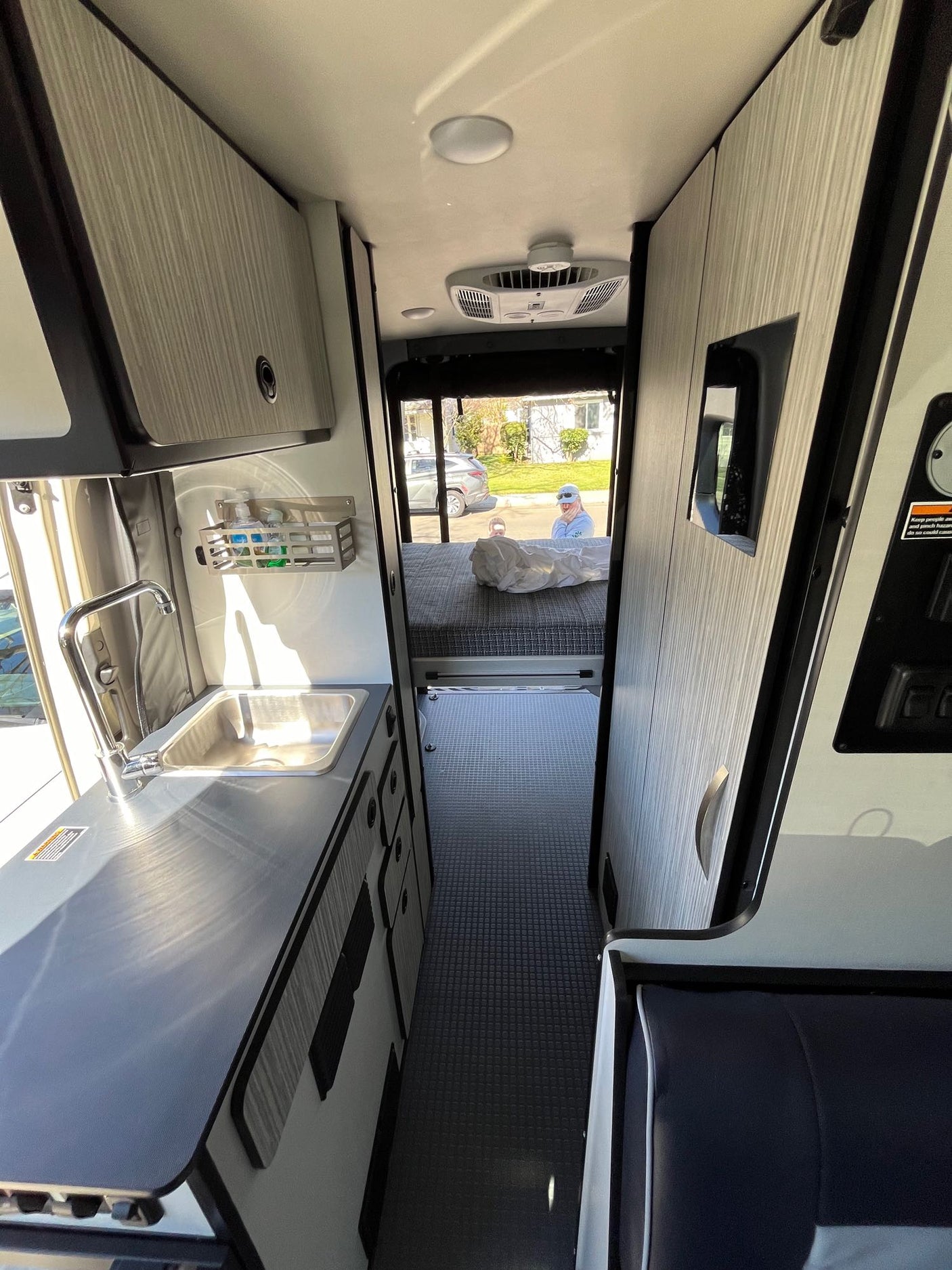 View inside an RV rental: a compact kitchen with a sink on the left, an aisle, and a bed at the rear. Functional camper van interior.