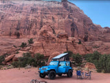 A blue Jeep, featuring a pop-top tent for an RV rental experience, is parked with camping chairs before grand red cliffs.