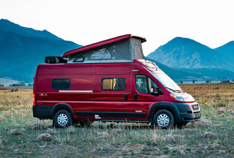 Red Winnebago Solis camper van with pop-top extended, parked in a scenic mountain field. Ideal for an RV rental adventure.