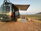 An adventure-ready RV rental camper van with an extended awning and outdoor setup for desert camping, complete with rug and fire pit.