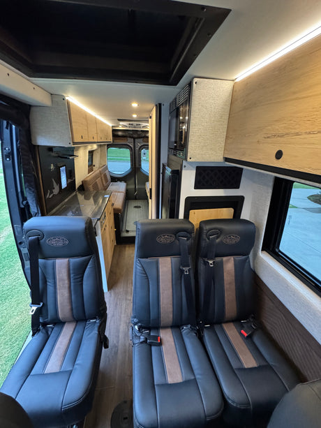 RV rental interior: black and brown passenger seats lead to a modern kitchen and dining area in this custom camper van.