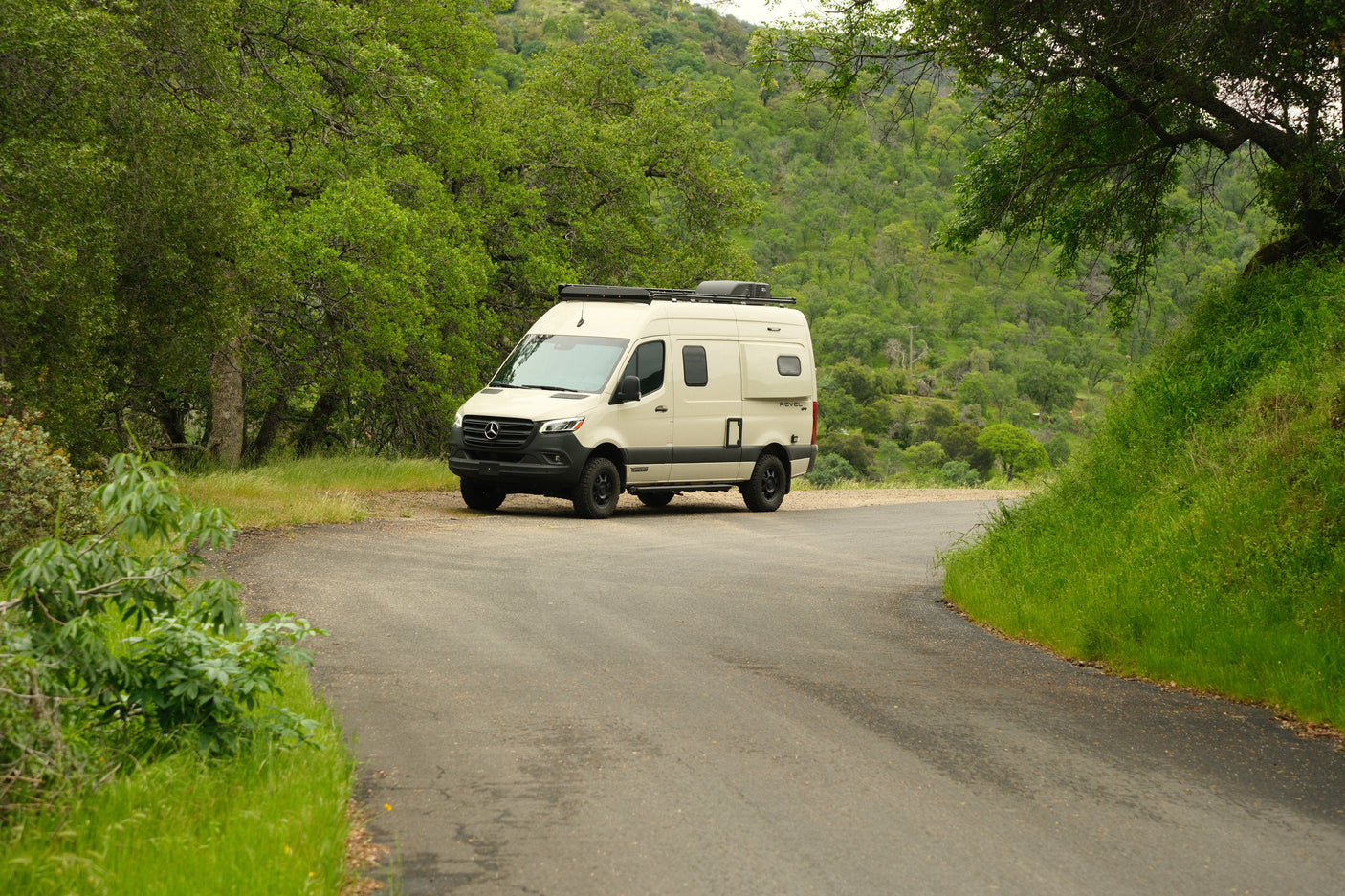 RV rental: A cream Winnebago Revel camper van is parked on a winding road amid lush green hills, ready for adventure.