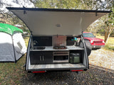 This RV rental teardrop camper's open rear reveals a full outdoor kitchen with stove, microwave, sink & cooler, at a campsite.