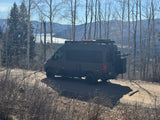 Dark adventure camper van, perfect for an rv rental, parked on a dirt road overlooking a scenic lake and mountains.