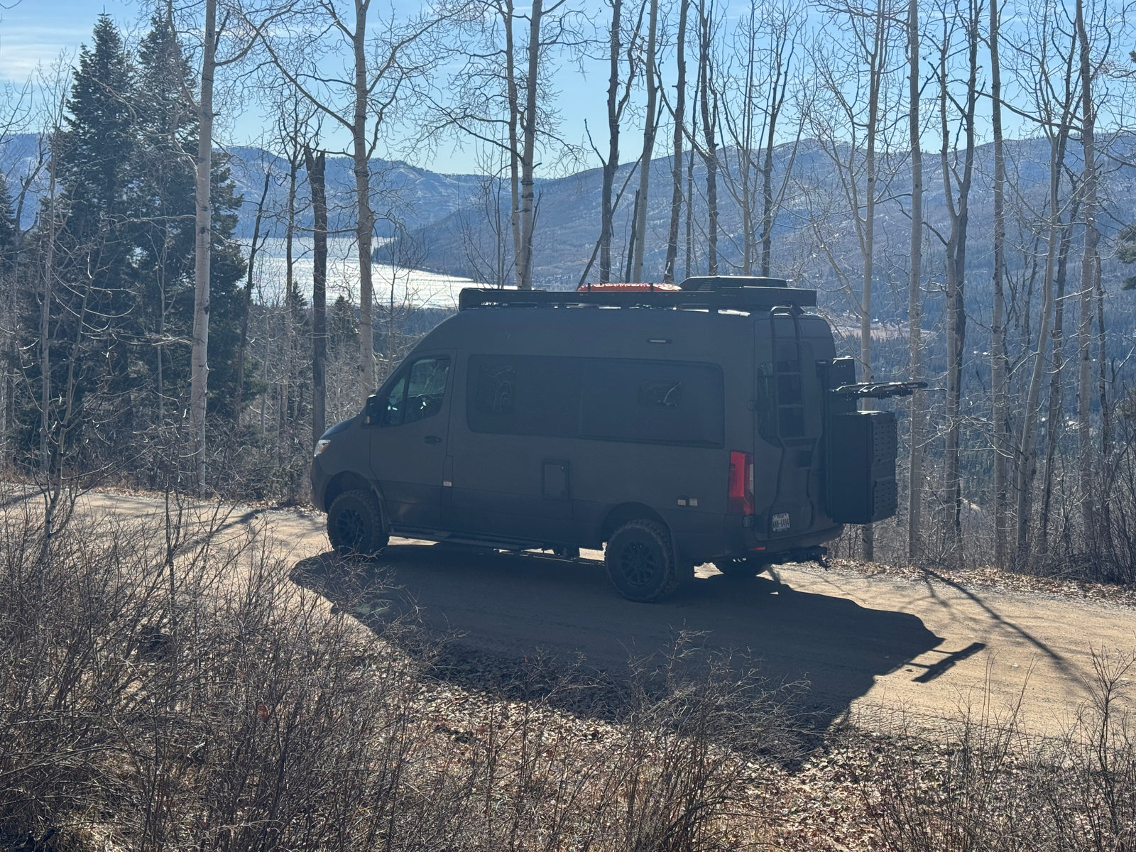 Dark adventure camper van, perfect for an rv rental, parked on a dirt road overlooking a scenic lake and mountains.