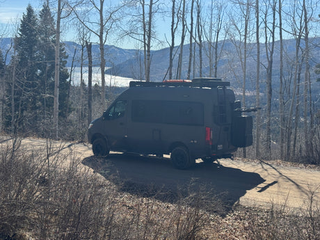 Dark adventure camper van, perfect for an rv rental, parked on a dirt road overlooking a scenic lake and mountains.