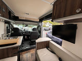 View of an rv rental interior: compact kitchen with sink, comfy seating, and TV. Driver's cab visible upfront.