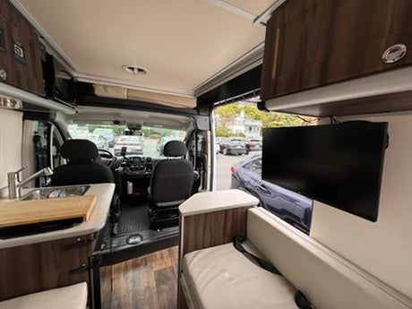 View of an rv rental interior: compact kitchen with sink, comfy seating, and TV. Driver's cab visible upfront.