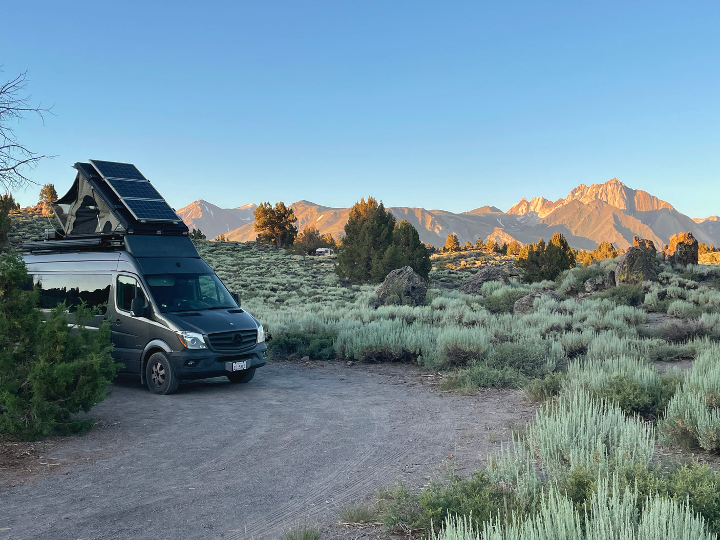 An RV rental camper van with solar panels is parked on a dirt patch amidst sagebrush, with sunlit mountains behind.