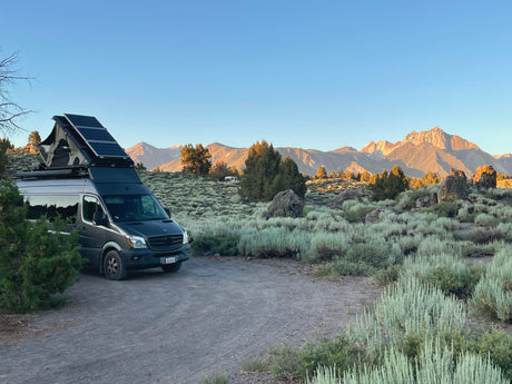 An RV rental camper van with solar panels is parked on a dirt patch amidst sagebrush, with sunlit mountains behind.