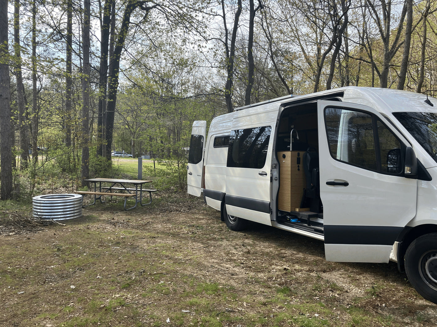 White rv rental camper van parked at a rustic campsite with a picnic table and fire pit, ideal for outdoor adventure.