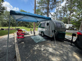 Perfect RV rental campsite setup! A sleek travel trailer, picnic table, and bikes under a canopy on a gravel spot.