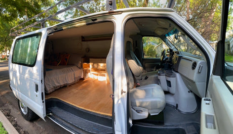Inside this spacious RV rental: a converted camper van with an open side door, showing a bed, wood floor, & front cabin.