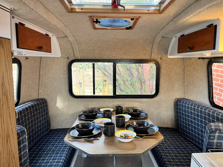 Cozy RV rental interior: dining area with a set table, blue plaid seats, windows, and overhead storage.