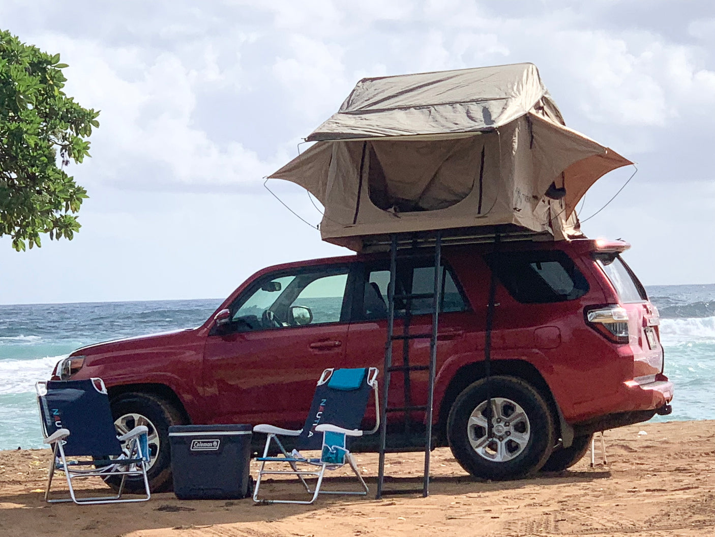 A red SUV with a rooftop tent, ladder, chairs, and cooler set up on a sandy beach. Great for an RV rental camping trip by the ocean.