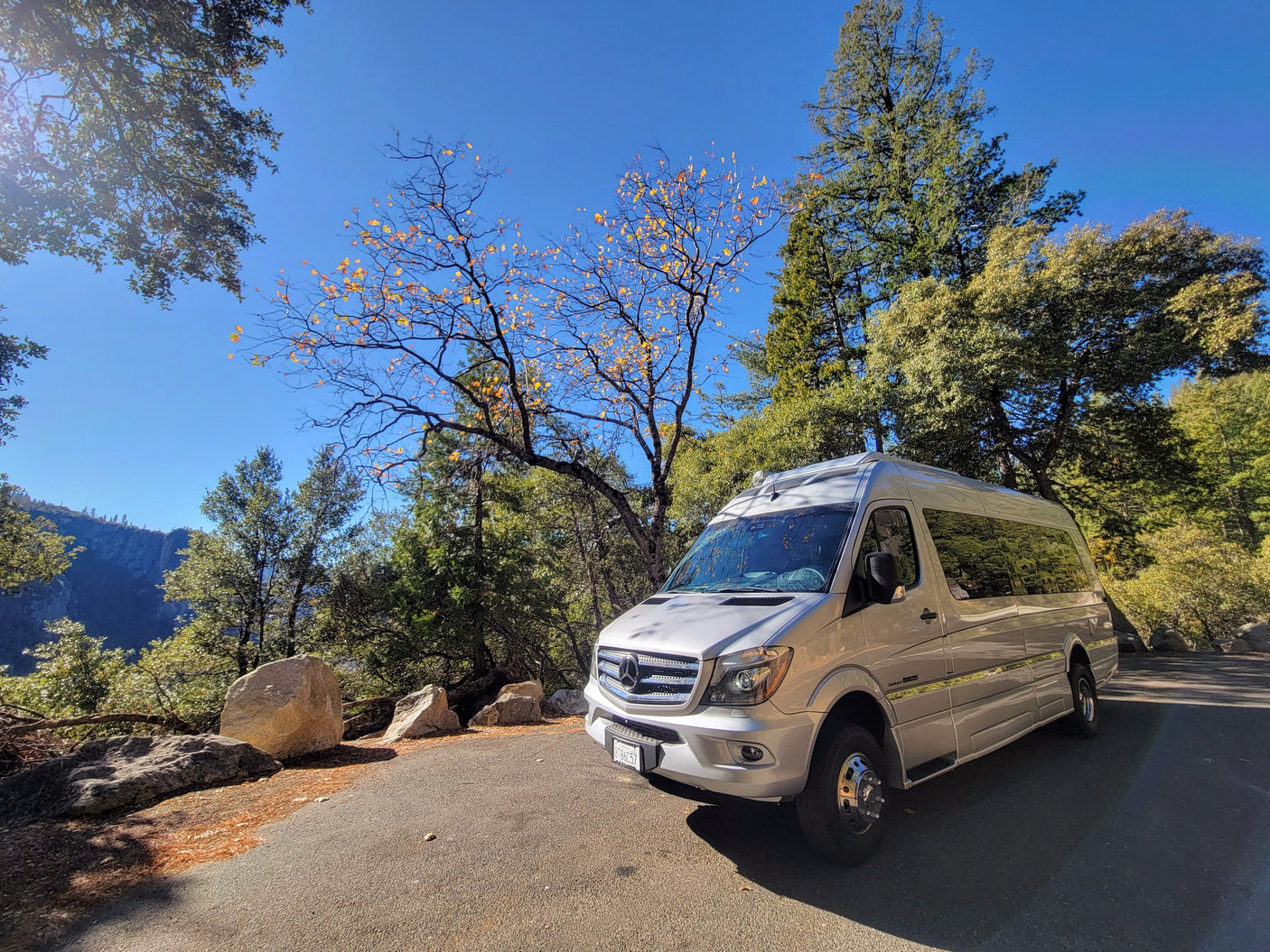 Silver Mercedes Sprinter RV rental on a sunny road. Scenic view with autumn trees & distant mountains.