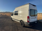 White Ford Transit RV rental, ready for adventure, parked on gravel in a scenic mountain setting under a clear blue sky.