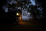 Twilight camping with a warm-lit RV rental van by a lake. String lights illuminate the dock, chairs, and trees.
