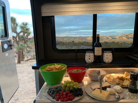 An RV rental's cozy setup: desert landscape, Joshua tree, and a delicious spread of wine, cheese, fruit, chips & salsa.