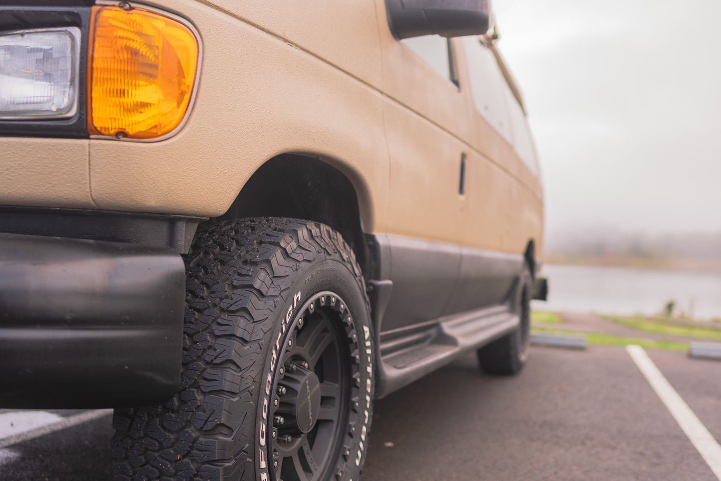 Close-up of a rugged tan adventure van with all-terrain tires, parked near a misty lake. Explore with an RV rental.