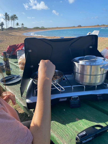 Outdoor cooking on a Coleman stove by a scenic beach, perfect with an RV rental. A person stirs food in a pan, pot nearby.