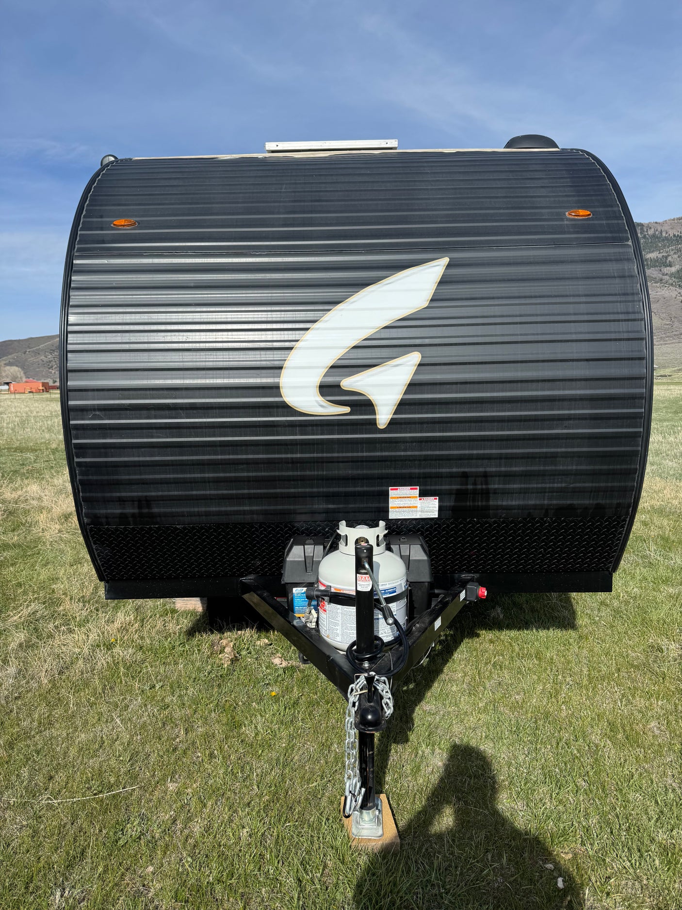 Black travel trailer (RV rental) with a white "G" logo, corrugated front, propane tank, and hitch, parked on green grass.