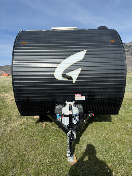 Black travel trailer (RV rental) with a white "G" logo, corrugated front, propane tank, and hitch, parked on green grass.