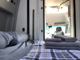 Cozy RV rental interior with a bed, plaid blanket, and stacked towels, looking towards the front cabin.