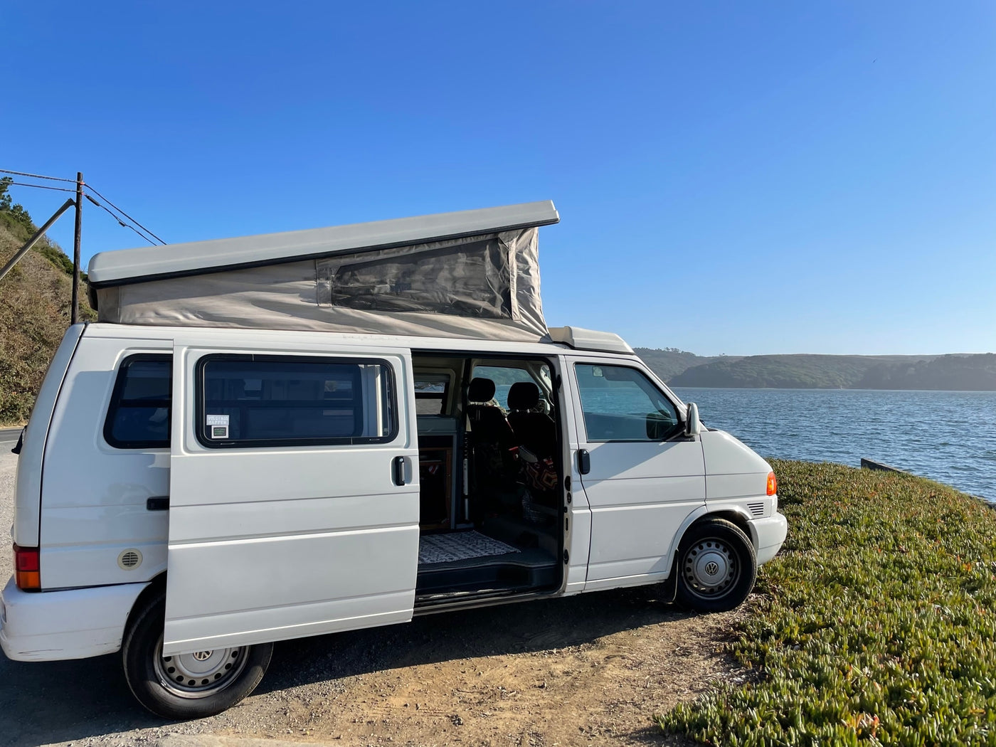 parked at a scenic coastal overlook among trees.|Cozy interior of an RV rental tent. Enjoy panoramic views of the sky and vibrant autumn trees from this comfortable sleeping space.|White pop-top VW camper van with open door — photo 2