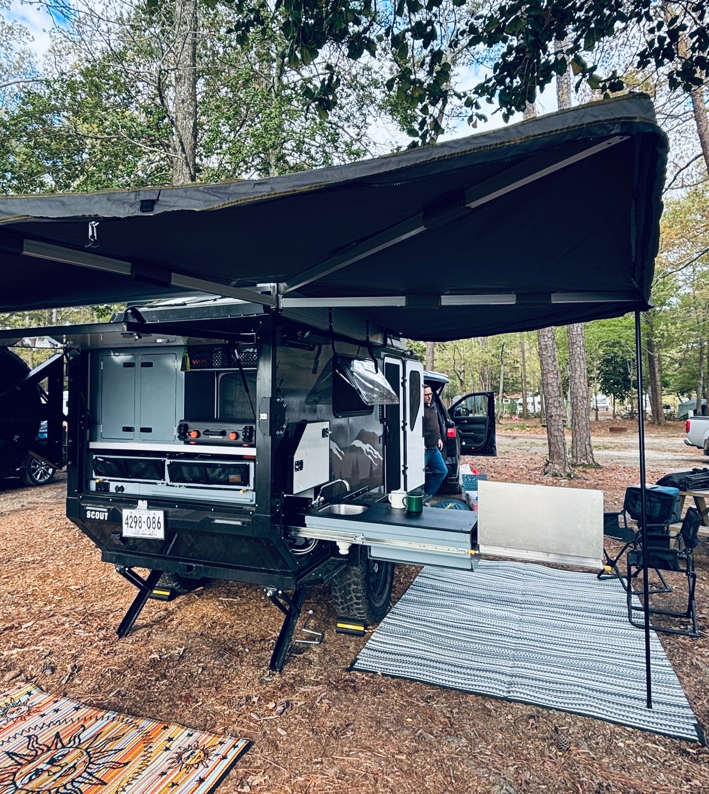 Enjoy this RV rental! An off-road camping trailer with an extended awning and full outdoor kitchen is set up at a wooded campsite.