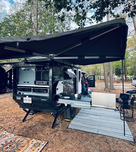 Enjoy this RV rental! An off-road camping trailer with an extended awning and full outdoor kitchen is set up at a wooded campsite.