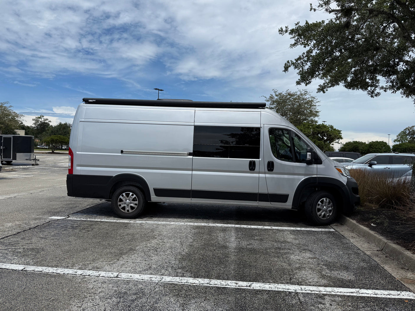 A silver RV rental camper van with a roof awning, parked in an asphalt lot under a partly cloudy sky.