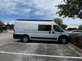 A silver RV rental camper van with a roof awning, parked in an asphalt lot under a partly cloudy sky.