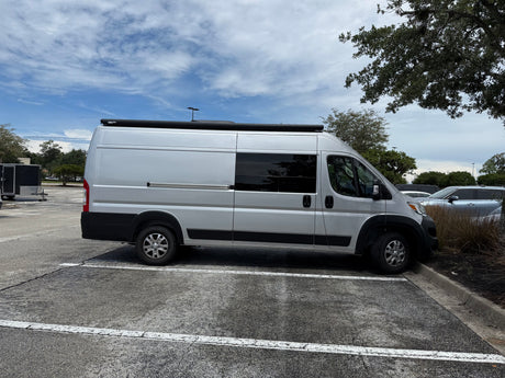 A silver RV rental camper van with a roof awning, parked in an asphalt lot under a partly cloudy sky.