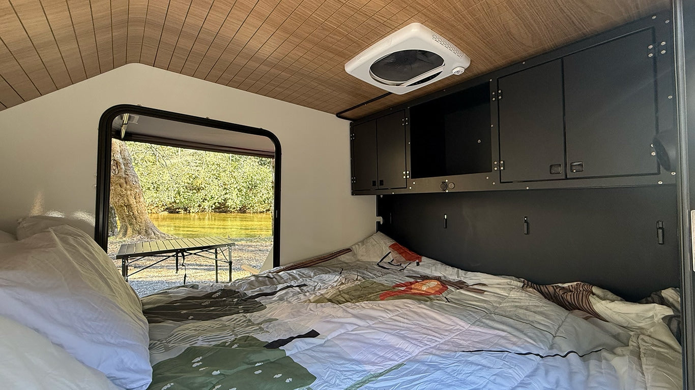 Cozy RV rental interior with bed, overhead storage, and a scenic view of a river/lake and trees outside the open door.