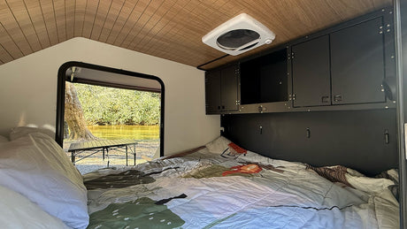 Cozy RV rental interior with bed, overhead storage, and a scenic view of a river/lake and trees outside the open door.