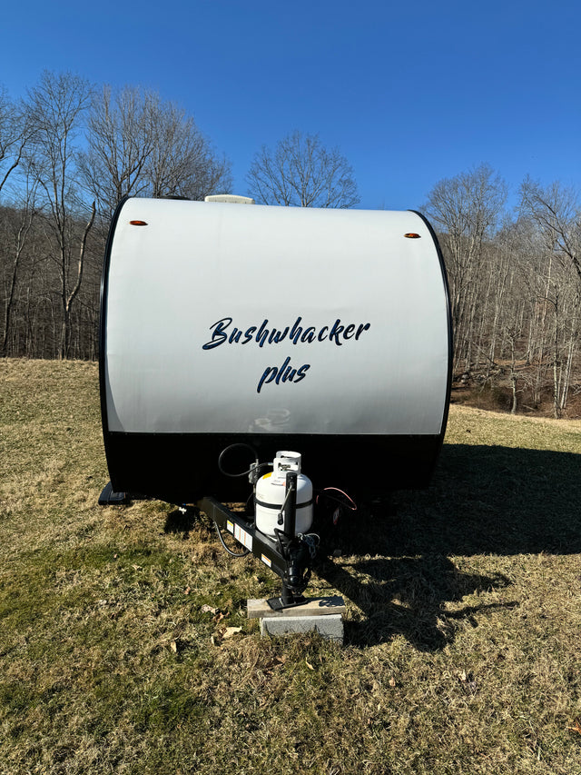 A white Bushwhacker Plus travel trailer, an ideal RV rental for adventures, sits under a clear blue sky with bare trees.