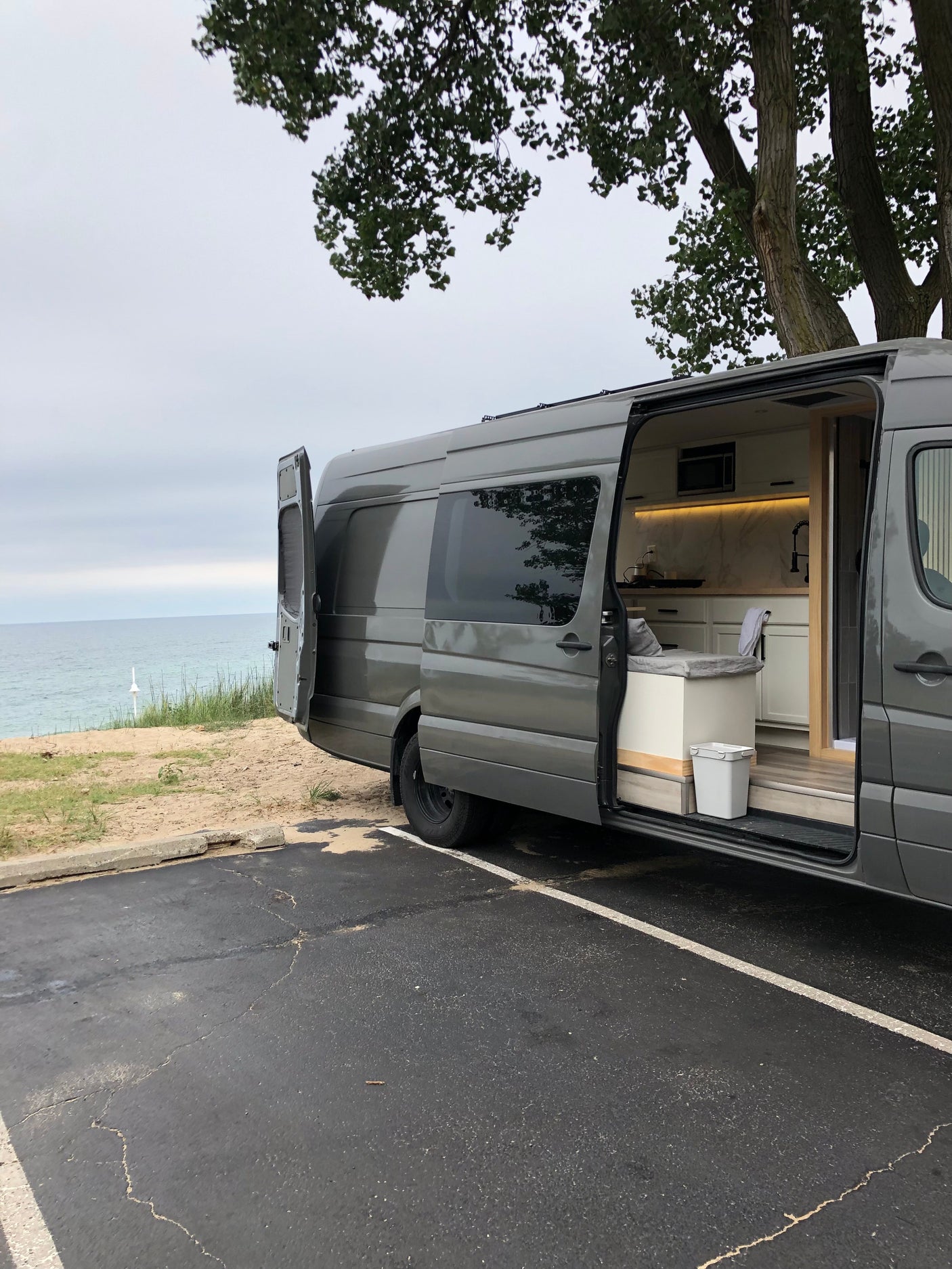 Grey rv rental van, open doors reveal a custom interior kitchen, parked by the water with trees overhead.