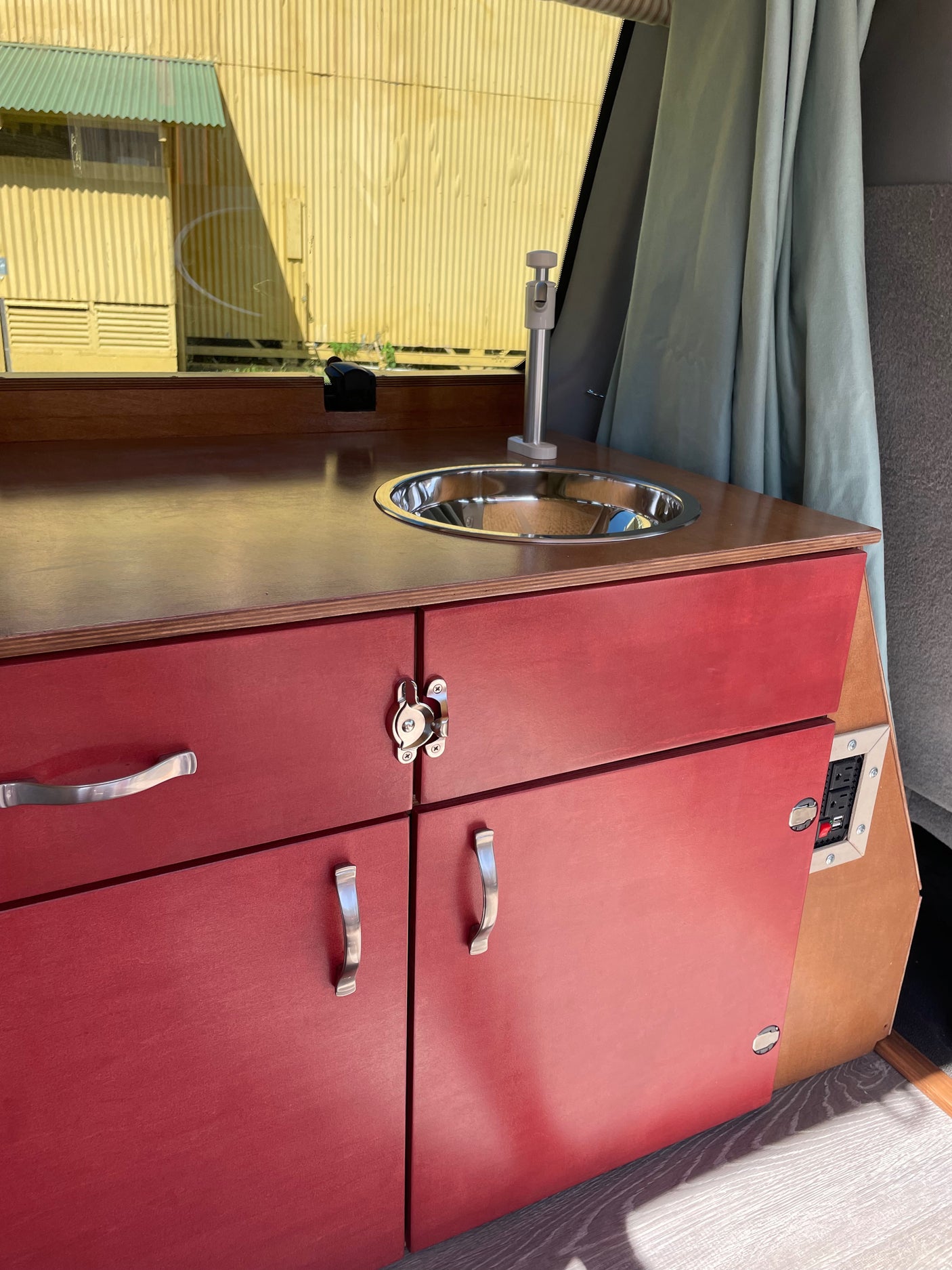 Compact kitchenette in an RV rental, featuring red cabinets, a stainless steel sink, and power outlets. Ideal for camper van journeys.